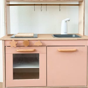 May include: A wooden toy kitchen in pink and natural wood tones. It has a microwave, oven, sink, and storage. Includes decorative wooden shapes and two clear cups with designs. A fun and engaging toy for children.