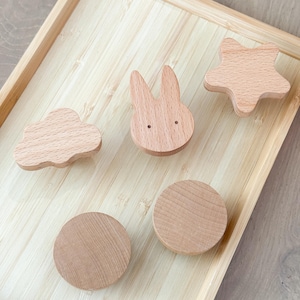 Puede incluir: Una colección de juguetes de madera, que incluye una nube, un conejito, una estrella y dos círculos, dispuestos sobre una bandeja de bambú. Los juguetes son de madera clara y tienen un acabado liso. El conejito tiene dos pequeños agujeros para los ojos.