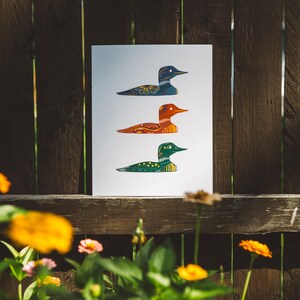 May include: A print of three stylized loon birds in a row. The top loon is blue, the middle loon is orange, and the bottom loon is green. Each loon has a patterned design.