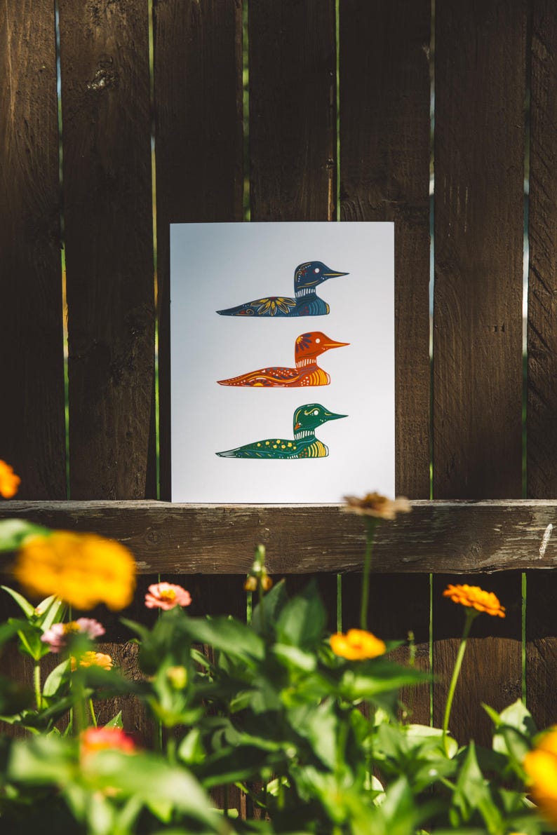 May include: A print of three stylized loon birds in a row. The top loon is blue, the middle loon is orange, and the bottom loon is green. Each loon has a patterned design.