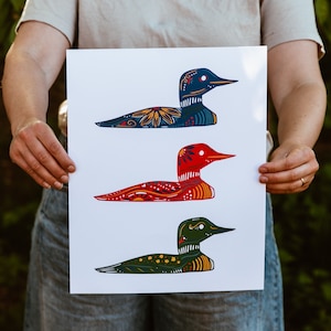 May include: A print featuring three stylized loon birds with colorful patterns. The birds are in a row, with the top bird facing right, the middle bird facing left, and the bottom bird facing right. The birds are all different colors, with the top bird being blue, the middle bird being red, and the bottom bird being green.