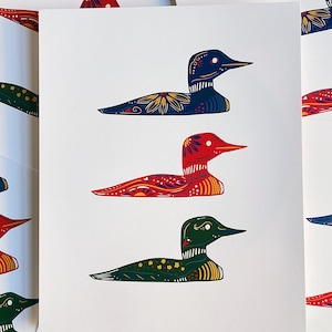 May include: A set of three colorful loon prints. The loons are depicted in a folk art style with intricate patterns and bright colors. The loons are in red, blue, and green.
