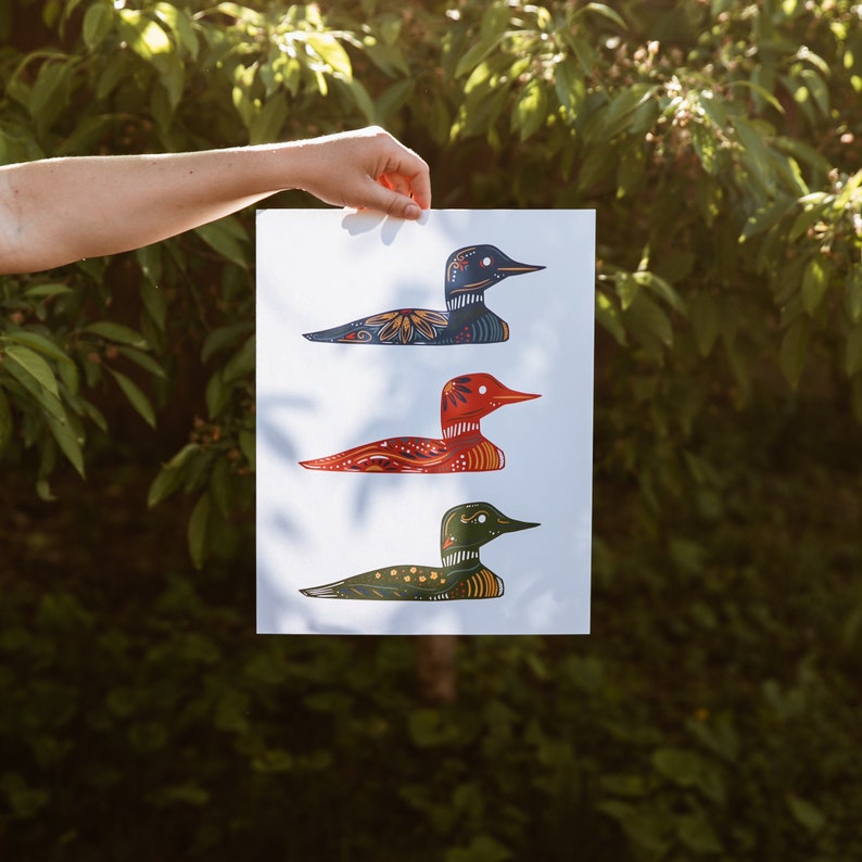 May include: A white sheet of paper with three colorful loon illustrations. The loons are in a row, with the top loon facing left, the middle loon facing right, and the bottom loon facing left. The loons are all different colors, with the top loon being blue and green, the middle loon being red and orange, and the bottom loon being green and black.