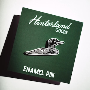 Puede incluir: Un pin de esmalte en blanco y negro con una ilustración detallada de un somorgujo. El pin está colocado sobre una tarjeta verde oscuro con las palabras "Hinterland GOODS" y "ENAMEL PIN" en blanco.