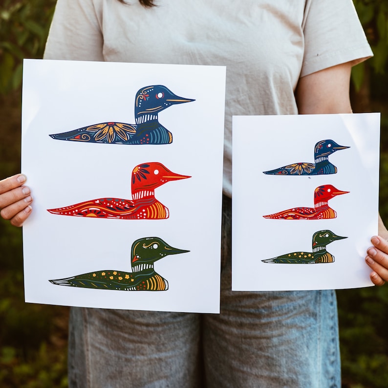 May include: Two prints of colorful loon illustrations. The top print features a blue loon with yellow and red details. The bottom print features a red loon with yellow and blue details. Both prints have a white background.