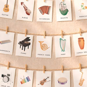 Puede incluir: Una colección de tarjetas ilustradas con varios instrumentos musicales, como una guitarra, un piano y un saxofón. Cada tarjeta está sujeta a una cuerda con una pinza de madera, creando una exhibición decorativa. Las tarjetas tienen los nombres de los instrumentos.