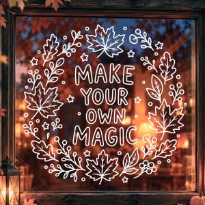 Puede incluir: Un adhesivo blanco para ventana que presenta una corona circular de hojas de otoño, bayas y pequeñas estrellas que rodean el texto "MAKE YOUR OWN MAGIC" en una fuente sans-serif clara, diseñado para decoración estacional en cristal.