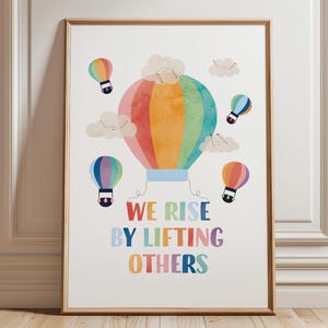Può includere: Una stampa artistica incorniciata con un design di mongolfiera colorata e la scritta "WE RISE BY LIFTING OTHERS". L'opera d'arte include un grande pallone dai colori dell'arcobaleno e diversi palloncini più piccoli, il tutto su sfondo bianco.