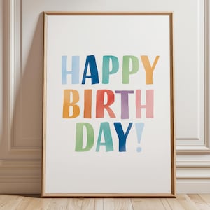 May include: A framed print with the words "HAPPY BIRTH DAY!" in colorful watercolor lettering. The letters are in shades of blue, green, orange, red, and purple. The frame is a light brown color.