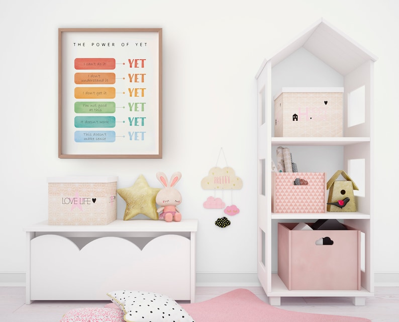 May include: A white framed motivational poster with the text "THE POWER OF YET" and a list of phrases with the word "YET" at the end of each phrase. The poster is hanging on a white wall. There is a white toy house shelf unit with pink storage boxes and a birdhouse on the top shelf. There is a white storage bench with a pink and white storage box on top of it. There is a pink and white rug on the floor.