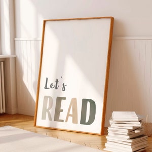 Peut inclure: Une impression encadrée avec le texte "Let's READ" dans un schéma de couleurs brun clair, beige et vert. L'impression est accrochée à un mur blanc avec une pile de livres au premier plan.