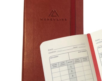 May include: A red leather-bound notebook with the words "MONEYLINE" embossed on the front cover. The notebook is open to a page with a scoring chart for a boxing match. The chart includes columns for round score, point deduct, and round score for both the red and blue corners.