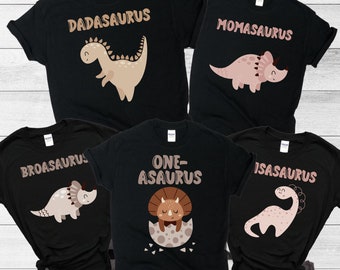 May include: Set of five black t-shirts with dinosaur graphics and text. The shirts read "Dadasaurus", "Momasaurus", "Broasaurus", "One-asaurus", and "Isasaurus".