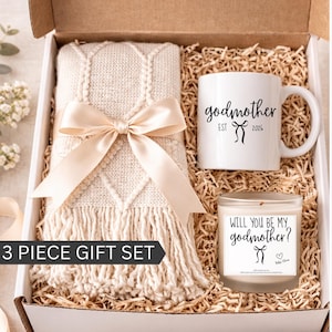 May include: A 3-piece gift set in a box, including a cream-colored blanket with a bow, a white mug with "godmother" text, and a candle. The mug and candle have black text. The blanket has a diamond pattern.
