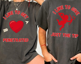 May include: Two dark gray t-shirts with red text and graphics. One shirt says "I LIKE TO GET PENETRATED" with a red heart pierced by an arrow. The other says "I LIKE TO GIVE JUST THE TIP" with a red Cupid.