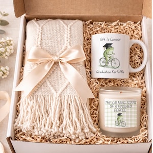 May include: A gift box with a cream-coloured throw blanket, diamond pattern, and fringe, tied with a satin bow. Includes a mug with a frog on a bicycle and text "Off To Commit" and a candle with text "The Calming Scent of a Finished Degree."