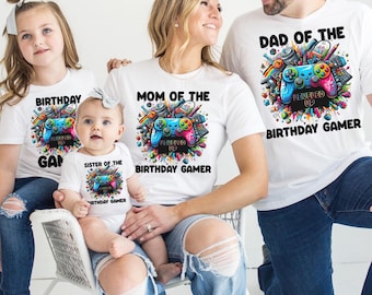 May include: A family of four wearing white t-shirts with a video game theme. The shirts feature a colorful illustration of a video game controller with the text "Birthday Gamer" and "Leveled Up". The shirts are available in different sizes for adults and children.