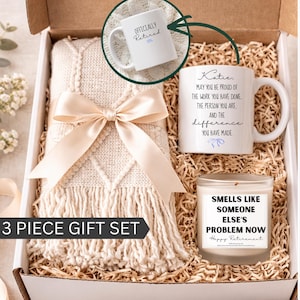 May include: A 3-piece gift set in a box, featuring a cream-colored throw blanket tied with a satin bow, two white mugs with retirement messages, and a candle. The mugs have text that reads "Officially Retired" and "Katie, May you be proud..." The candle reads "Smells like someone else's problem now."
