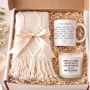 May include: A gift box containing a cream-colored knitted throw blanket tied with a satin ribbon, a white mug with text, and a scented candle. The mug reads "a new chapter" with an inspirational quote. The candle has text that reads "Smells like your next chapter is just beginning."