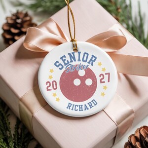 May include: A round white ceramic ornament featuring a red bowling ball graphic in the center. The text reads SENIOR, Striker, RICHARD, and the numbers 20 and 27, surrounded by small yellow stars. It hangs from a thin gold string.