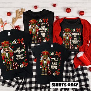 May include: Three black t-shirts featuring a colorful nutcracker graphic, red bows, and white flowers. The shirts display the text: "This nutcracker is totally cracked," "Deez nuts roasting on an open fire," and "Y'all are nuts" with holiday accents.