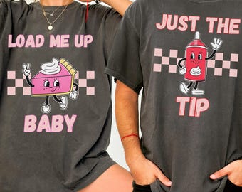 May include: Two dark gray t-shirts with matching graphics. One shirt says "LOAD ME UP BABY" with a cartoon pie slice. The other says "JUST THE TIP" with a cartoon ketchup bottle. Both have pink and white checkered accents.