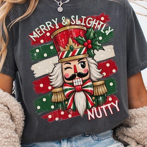 May include: A charcoal gray t-shirt featuring a graphic of a winking nutcracker wearing a red and green hat with holly. The design includes the text MERRY & SLIGHTLY NUTTY in white lettering over a background of red and green horizontal stripes.