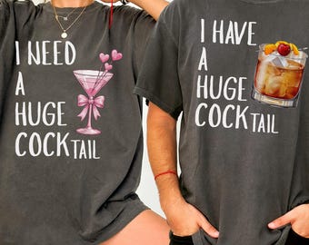 May include: Two charcoal gray t-shirts with white text and cocktail graphics. One shirt says "I NEED A HUGE COCKTAIL" with a pink martini glass. The other says "I HAVE A HUGE COCKTAIL" with a cocktail.