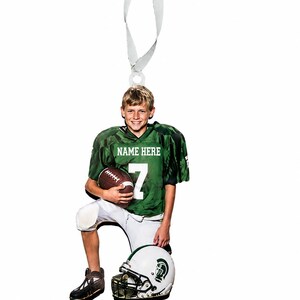 May include: A flat, die-cut ornament featuring a football player in a green jersey with the text NAME HERE and the number 7, white pants, and dark cleats. The player holds a brown football and rests a hand on a white and green football helmet.