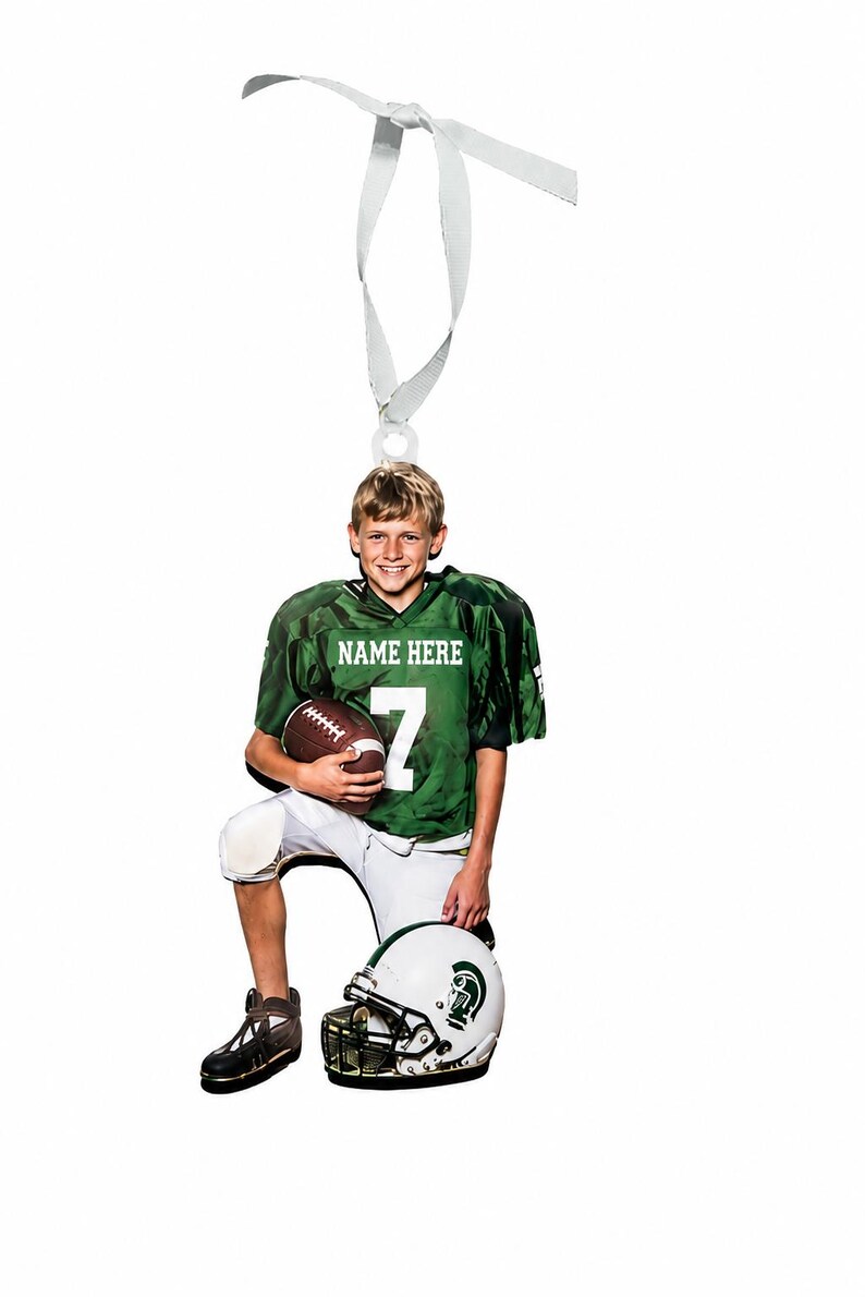 May include: A flat, die-cut ornament featuring a football player in a green jersey with the text NAME HERE and the number 7, white pants, and dark cleats. The player holds a brown football and rests a hand on a white and green football helmet.