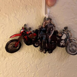May include: A custom-shaped hanging ornament featuring a photographic print of two people standing between two motorcycles. The left motorcycle is red, and the right motorcycle is white and black. The ornament is suspended by a white ribbon.
