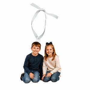 May include: Two children kneeling side by side, one wearing a navy blue long-sleeved shirt and blue jeans, the other in a cream-colored long-sleeved top and blue jeans, with a white ribbon loop positioned above their heads against a white background.
