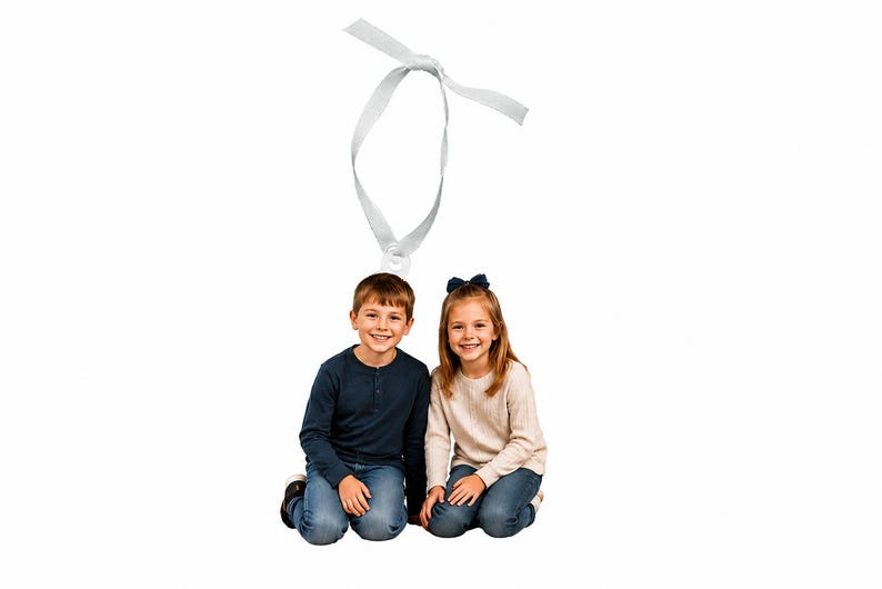 May include: Two children kneeling side by side, one wearing a navy blue long-sleeved shirt and blue jeans, the other in a cream-colored long-sleeved top and blue jeans, with a white ribbon loop positioned above their heads against a white background.