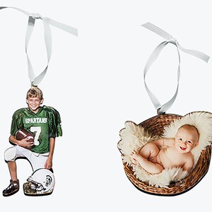 May include: Two custom photo ornaments with white ribbon hangers. One ornament features a child in a green Spartans football jersey holding a football with a helmet nearby. The other ornament shows a baby lying in a brown woven basket with fur.