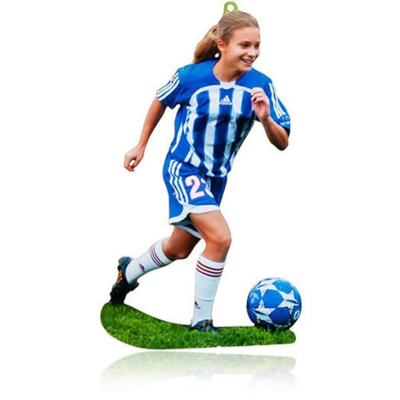 May include: A flat, die-cut ornament featuring a soccer player in a blue and white striped jersey and matching shorts with the number 23. The player is running on a patch of green grass while dribbling a blue and white soccer ball with star patterns.