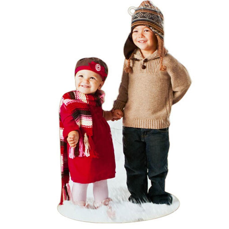 May include: A cardboard cutout of two children standing together. One child wears a red dress, a red and white striped scarf, and a red headband. The other child wears a tan sweater, dark blue jeans, and a brown patterned knit hat with earflaps.