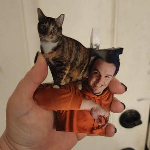 May include: A custom acrylic ornament featuring a photographic print of a person wearing an orange hoodie with a brown and black tabby cat perched on their shoulder. The ornament includes a white plastic hanging loop at the top for display.
