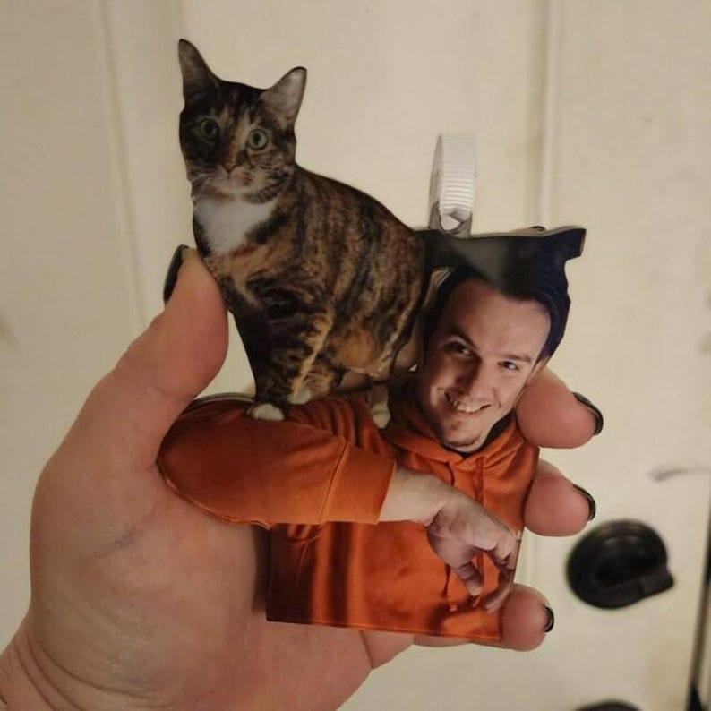 May include: A custom acrylic ornament featuring a photographic print of a person wearing an orange hoodie with a brown and black tabby cat perched on their shoulder. The ornament includes a white plastic hanging loop at the top for display.
