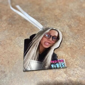 May include: A custom acrylic ornament featuring a photograph of a person with long blonde hair, wearing black-rimmed glasses, and sticking their tongue out. The ornament includes a white ribbon for hanging and the text POSITIVE VIBES in colorful letters.