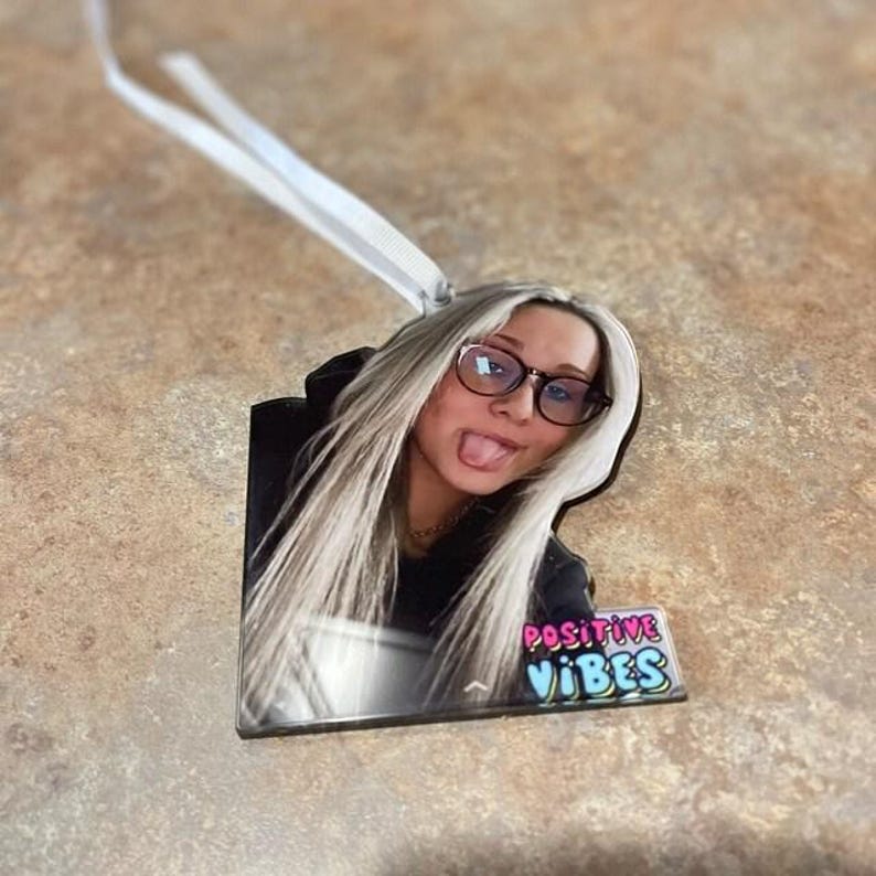 May include: A custom acrylic ornament featuring a photograph of a person with long blonde hair, wearing black-rimmed glasses, and sticking their tongue out. The ornament includes a white ribbon for hanging and the text POSITIVE VIBES in colorful letters.