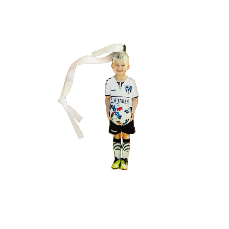 May include: A flat, die-cut ornament featuring a child in a white and black soccer uniform with the text "TOTTENHAM" on the shirt, holding a soccer ball with a blue and red star pattern. A white ribbon is attached to the top for hanging purposes.
