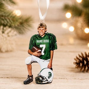 May include: A flat hanging ornament featuring a young football player in a green jersey with the text NAME HERE and the number 7, holding a brown football. The player wears white pants and kneels beside a white helmet with a green logo.