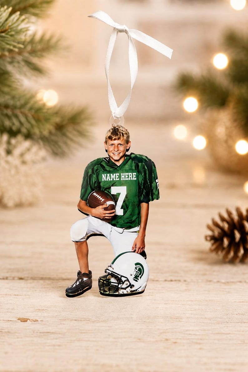 May include: A flat hanging ornament featuring a young football player in a green jersey with the text NAME HERE and the number 7, holding a brown football. The player wears white pants and kneels beside a white helmet with a green logo.