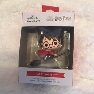 May include: A Hallmark Ornaments Harry Potter Christmas tree ornament in its original packaging. The figure depicts Harry Potter with round glasses and a lightning bolt scar, wearing a dark blue robe and riding a brown broomstick with black bristles.