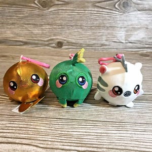 May include: Three plush toys with large eyes and pink cheeks. One is gold, one is green, and one is white with gray stripes. Each has a pink loop attached. The toys are round and have a cute, cartoonish design.