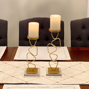 May include: Two gold metal candle holders with geometric designs. Each holder has a white pillar candle on top. The candle holders are on a white table runner with a diamond pattern.