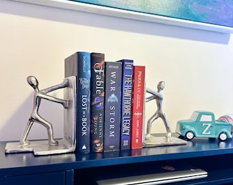 May include: Silver bookends shaped like stick figures holding up a row of five books with titles including "Fable", "War Storm", "The Hawthorne Legacy", and "Lost in a Book".