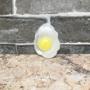 May include: A white and yellow soap shaped like a fried egg. The soap is on a brown and beige textured surface.