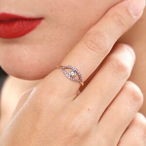 May include: A gold ring with a small white stone in the center, surrounded by a circle of purple stones. The ring is shaped like an eye.