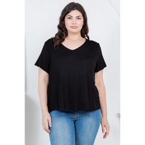 Op de afbeelding: Een vrouw draagt een zwart V-hals T-shirt en een blauwe spijkerbroek. De top met korte mouwen heeft een losse pasvorm. De jeans is licht gewassen denim. De achtergrond is een effen witte muur.